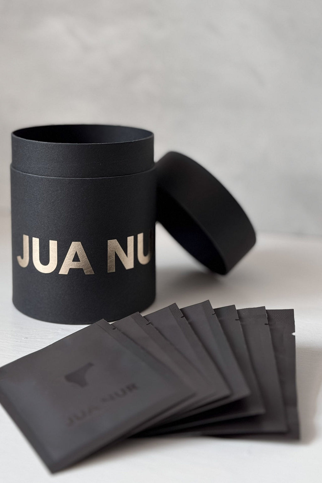Black cylindrical container with 'JUA NUR' branding, accompanied by black packets on a light gray background.