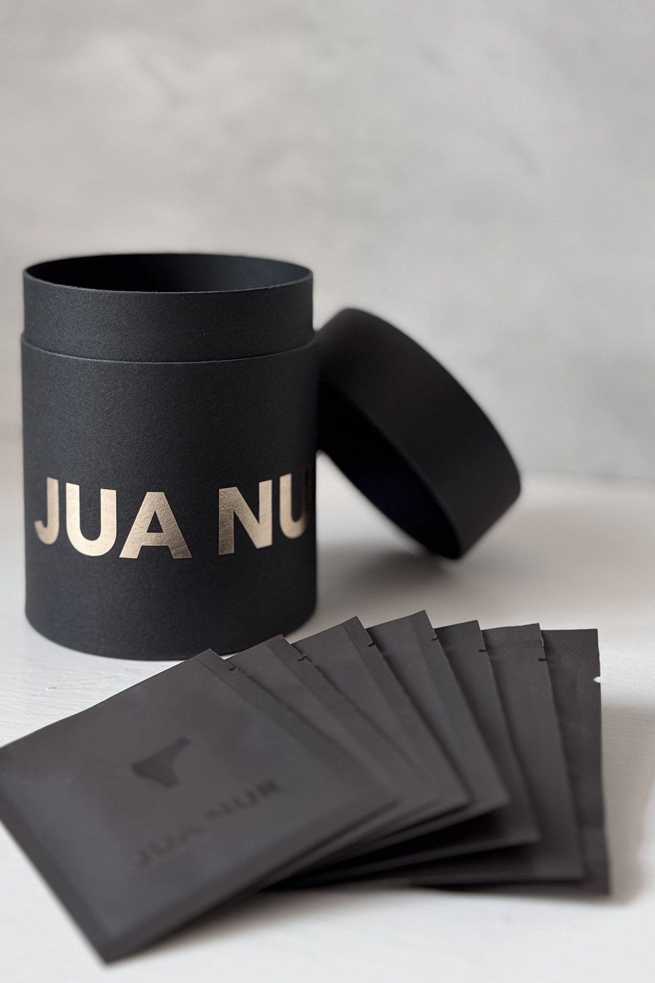 Black cylindrical container with 'JUA NUR' branding, accompanied by black packets on a light gray background.